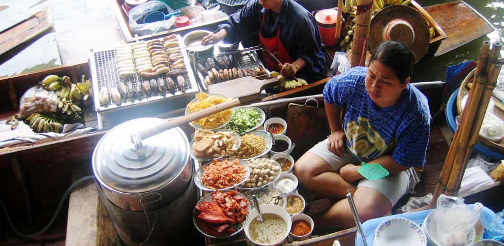 Thai vendor on a wooden boat at Floating Market Bangkok displaying and preparing fresh grilled foods, fruits, and dishes